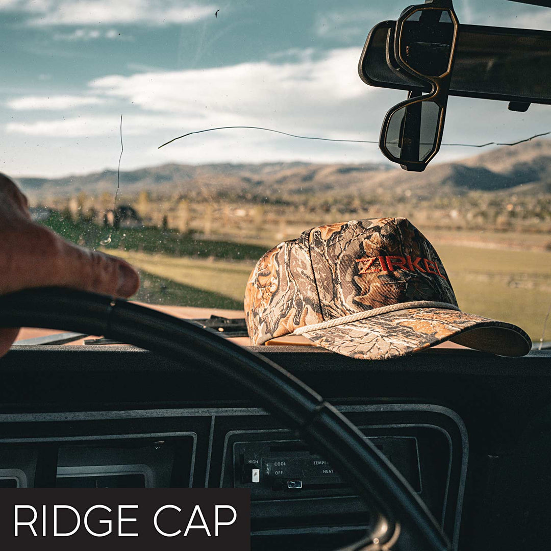 Camouflage Zirkel Ridge Cap on a car dashboard with a scenic view outside