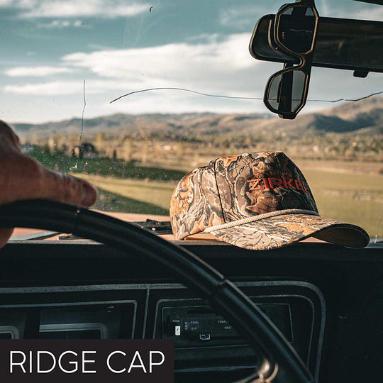 Camouflage Zirkel Ridge Cap on a car dashboard with a scenic view outside